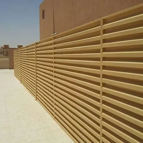 ساتر خشبي بخطوط أفقية يمتد بمحاذاة ممر خرساني بجانب مبنى بسيط في مدينة أبها، تحت سماء صافية، يُضفي لمسة طبيعية دافئة على المساحة الخارجية