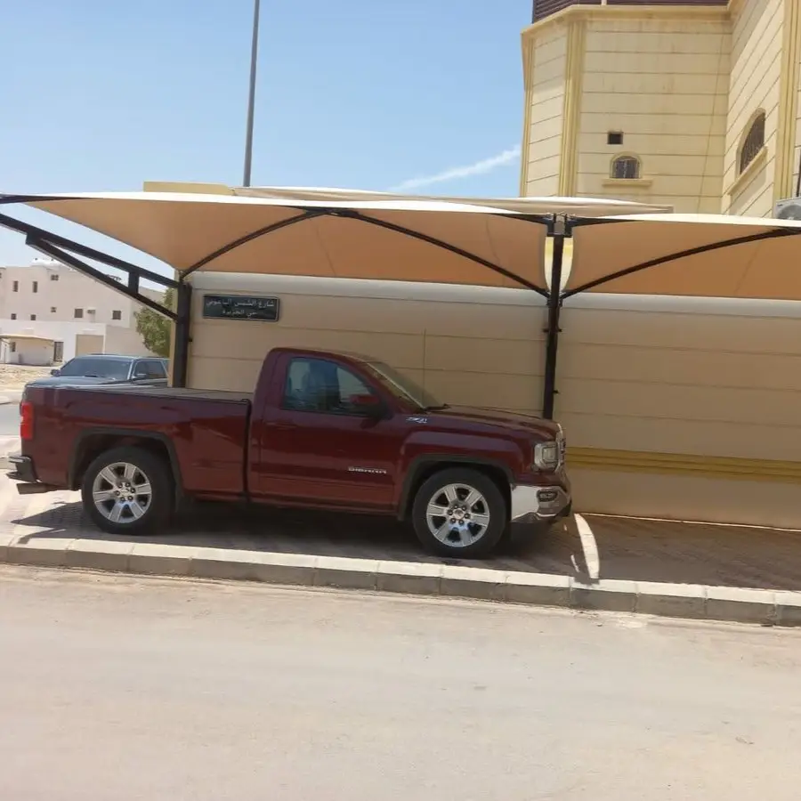 شاحنة بيك أب حمراء من نوع GMC Sierra متوقفة تحت مظلة قماشية بلون بيج، مدعومة بهيكل معدني أسود، بجانب مبنى بلون أصفر فاتح يحمل لافتة مكتوب عليها "مواقف السيارات للموظفين فقط".