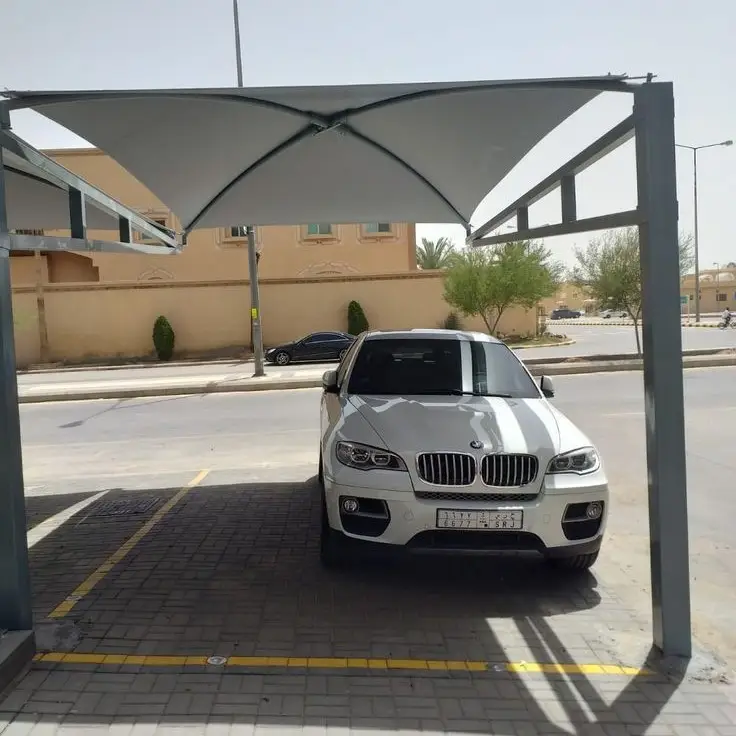 سيارة بي إم دبليو بيضاء متوقفة تحت مظلة قماشية حديثة مدعومة بهيكل معدني، في موقف سيارات محدد بخطوط صفراء، وخلفها شارع وأشجار ومبنى بلون بيج.
